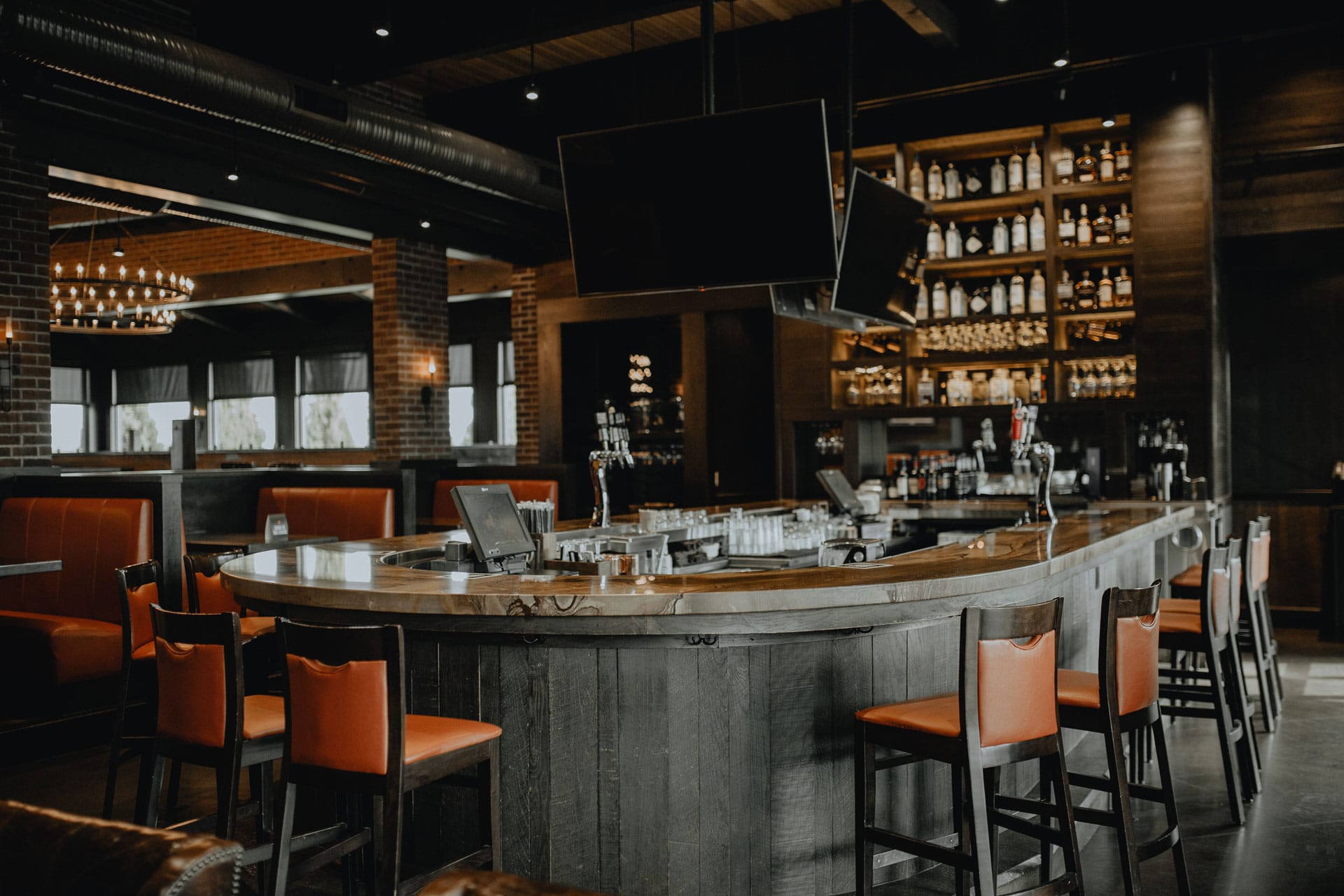 Curved full-service bar featuring high-backed orange chairs, overhead TVs, and illuminated liquor shelves.