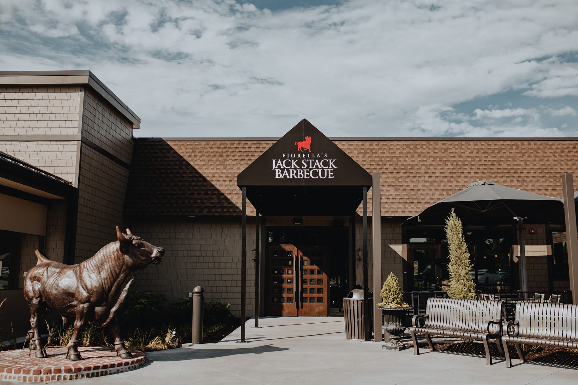 Exterior view of the restaurant entrance with a black Jack Stack Barbecue canopy, wooden doors, outdoor seating benches, and a bronze bull sculpture.
